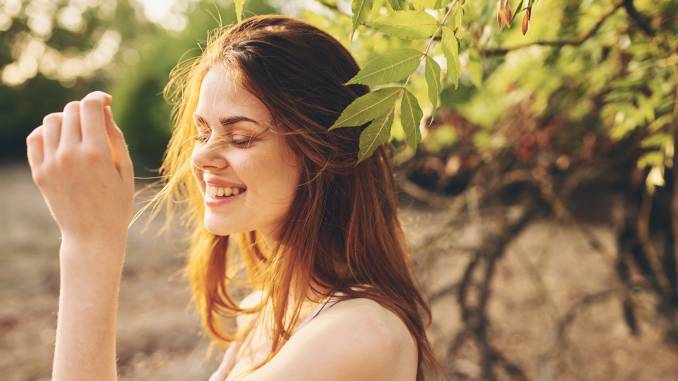 Jeune femme blanche, aux cheveux châtains, face au soleil, le sourire aux lèvres, dans un cadre de nature, de verdure. Le PABA (ex vitamine B10) jouerait-il un rôle dans la santé de sa peau et de ses cheveux ?